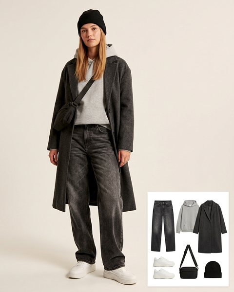 Woman wearing washed black baggy jeans with a grey hoodie, a long charcoal overcoat, chunky white sneakers, a black crossbody bag and a black beanie