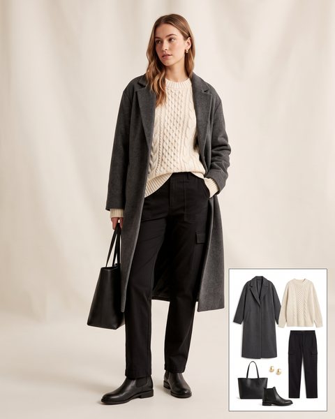 Woman wearing black straight-leg cargo pants with a cream cable-knit sweater, a long charcoal wool coat, black Chelsea boots and a black leather tote bag