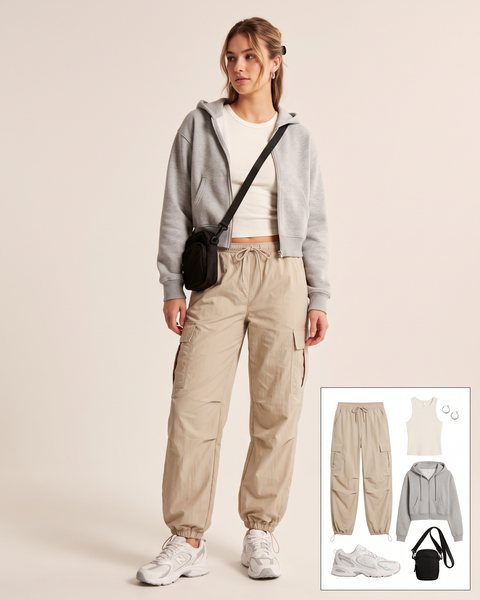 Woman wearing stone beige parachute cargo pants with an ivory ribbed tank, a cropped grey zip-up hoodie, chunky white sneakers and a small black crossbody bag