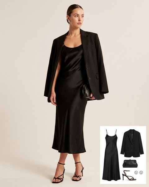 Woman wearing a black satin slip midi dress with an oversized black blazer on her shoulders, black strappy heels and a black clutch