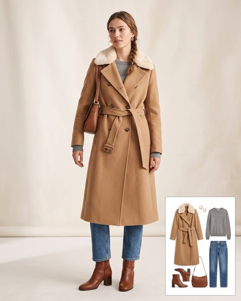 Woman wearing a camel wool belted coat with a cream faux fur collar over a grey sweater, straight jeans, cognac ankle boots and a cognac shoulder bag