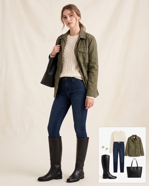 Woman wearing flat black leather riding boots with dark-wash skinny jeans tucked in, a cream cable-knit sweater, an olive green field jacket and a black leather tote