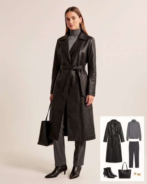 Woman wearing a long black leather trench coat with a grey turtleneck, charcoal tailored trousers, black kitten-heel ankle boots and a black leather tote bag
