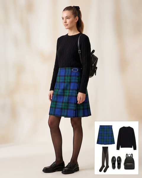 Woman wearing a royal blue and green tartan wrap kilt skirt with a black cable-knit sweater, sheer black tights, chunky black loafers and a small black backpack