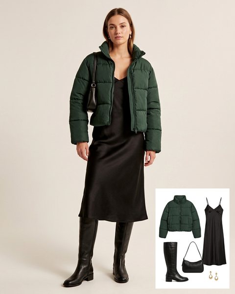 Woman wearing a dark green cropped puffer jacket over a black satin slip dress with black knee-high boots and a black shoulder bag