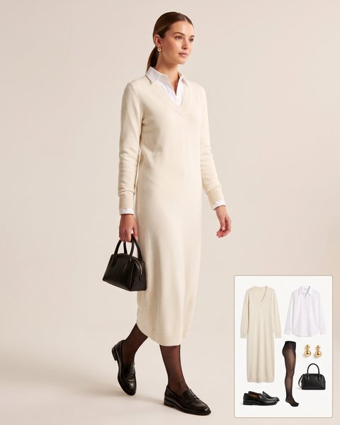 Woman wearing a cream V-neck knit midi dress over a white collared shirt with sheer black tights, black penny loafers and a small black top-handle bag