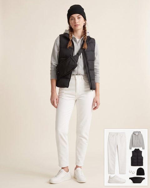 Woman wearing white mom jeans with a grey zip-up hoodie, a black puffer vest, white leather sneakers, a black belt bag, a black beanie and silver hoop earrings